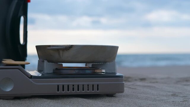 Portable camping stove cooking on sandy beach by the ocean. Portable gas stove holding empty cooking pot on sandy beach with rolling ocean waves, representing outdoor culinary preparation