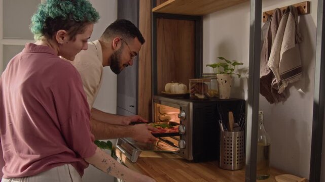 Tilt up view of young man putting baking sheet with pizza into electric oven and hugging short-haired girlfriend