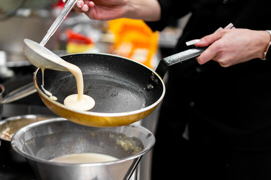 Hands pouring liquid batter into a hot frying pan to cook a thin pancake or crepe, with a mixing bowl in the foreground