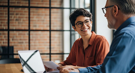 Older man and young woman collaborating over digital tablet in modern office setting with large windows