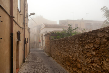 Narrow cobblestone street in Enna, Sicily, Italy, lined with old stone buildings and shrouded in morning fog, creating a mysterious atmosphere in the historic town