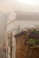 Foggy street view in Enna, Sicily, featuring traditional architecture, stone walls, and a vintage street lamp illuminating the narrow alleyway