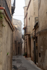 Narrow cobblestone alleyway in Enna, Sicily, featuring weathered stone buildings with balconies and potted plants, creating a historic urban atmosphere