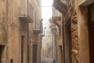 Narrow cobblestone street in Enna, Sicily, Italy, lined with rustic buildings featuring balconies and stone walls, shrouded in morning fog creating a serene atmosphere