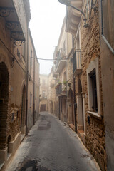 Narrow cobblestone street in Enna, Sicily, Italy, flanked by historic stone buildings with balconies and fog creating a serene atmosphere in the early morning light