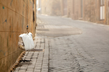 Hanging white garbage bag secured to a wall on a cobblestone street in Enna, Sicily, Italy, with blurred buildings and fog in the background