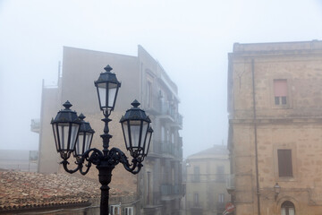 Vintage street lamp in foggy Enna, Sicily, highlighting historic architecture and buildings with a mysterious ambiance in the background