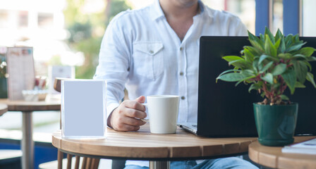 Fototapeta premium A white coffee mug beside a laptop on a wooden café table, with a person softly out of focus in the background, creating a calm modern workspace and lifestyle atmosphere.