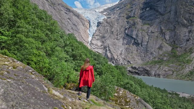 Woman in red jacket admiring Briksdalsbreen landscape in Norway. Mountain cliffs, green forest and glacier create a serene summer travel atmosphere.