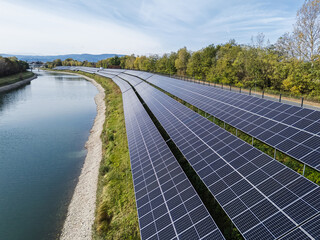 Aerial view shows rows of solar panels by the river with trees in the background under clear skies.