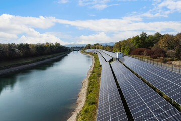Solar panels line the bank of a river under a clear sky with trees and hills in the background.