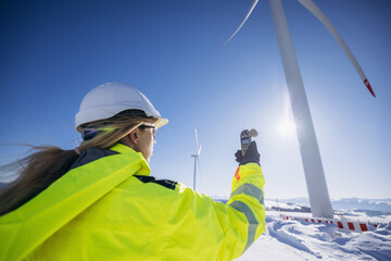 Female engineer standing in large field energy windmills and using meteorological station to measure the wind speed. Wind turbine station. Clean energy.