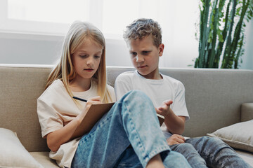 Obraz premium Two focused children sit on sofa indoors, girl writing on clipboard while boy explains with hand gestures. Concept of education, teamwork, learning, communication, and childhood development.