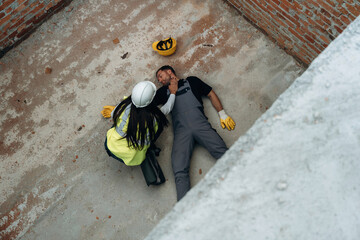 Fototapeta premium Top view. Man, construction worker fell from big height, lying down without consciousness, accident, woman is checking