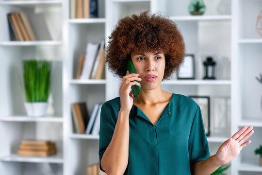 Young serious mixed race office worker female at workplace holding mobile phone makes business call listens client claims feels displeased annoyed and anxious, problems at work concept
