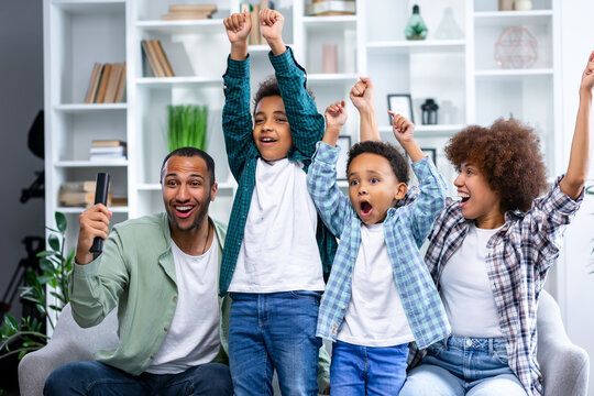 Low angle front view of four members family at home sitting on a sofa in the living room, watching TV and all cheering and rasing fists in victory
