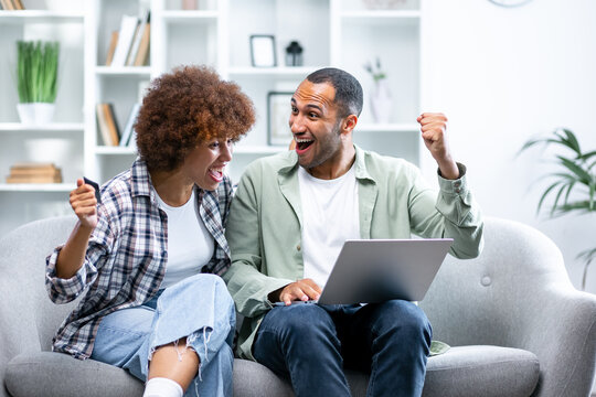 Joyful sincere laughing young african american family couple celebrating internet success, getting online lottery giveaway gambling auction win notification, feeling excited looking at laptop screen.