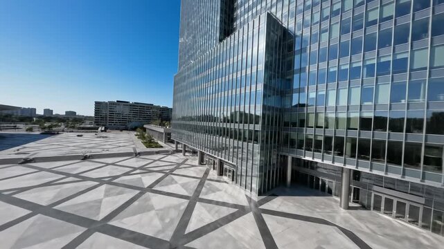 Sweeping aerial drone shot panning over a vast geometric plaza stretching towards the towering glass facade of a contemporary high-rise building geometric, highrise, openarea
