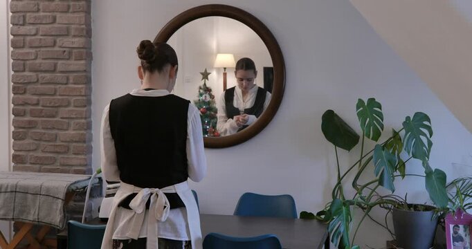 A young woman wearing period clothing and an apron is standing with her back to the camera, admiring her reflection in a round mirror and adjusting her earrings.