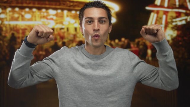 Young man enjoys lively outdoor night fair with blurred lights and crowd in background, capturing joyful atmosphere with confident expression and raised arms in casual sweater.