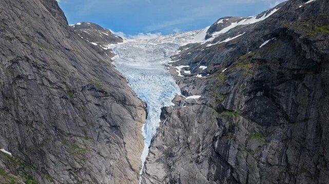 Briksdalsbreen glacier flowing through steep rocky gorge in Norway. Aerial drone view shows blue ice between towering cliffs under clear summer skies.