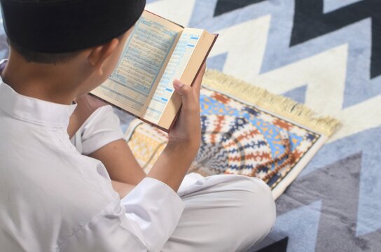 Overhead view of a muslim boy reading the Quran on a blue prayer mat.
