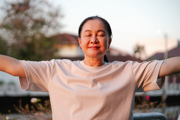 Older Asian woman standing with eyes closed and arms open during sunrise or sunset. Mindfulness, meditation, peaceful wellness and healthy lifestyle concept.