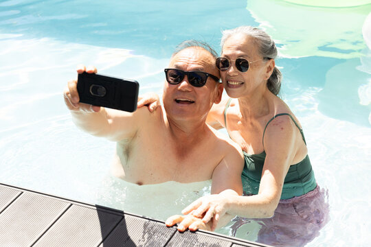 Mature Asian couple posing for selfie in clear pool by deck in swimsuits holding smartphone