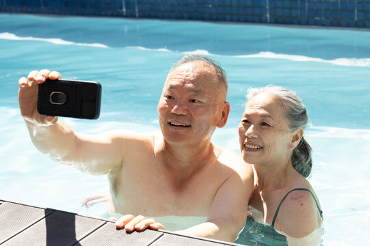 Senior Asian couple smiling, taking selfie in pool at edge, in swimwear holding black smartphone