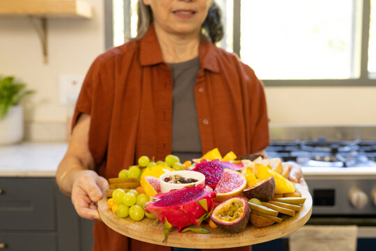 Round wooden platter is showcasing dragonfruit passionfruit grapes mango on sunlit kitchen counter