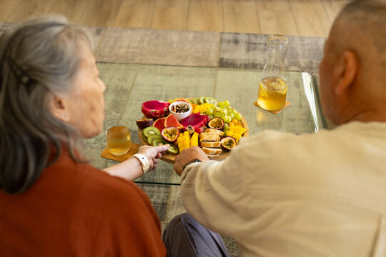 Senior couple sitting at glass coffee table reaching for shared dragon fruit platter and glasses