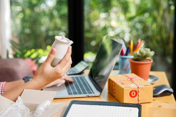 Close up of online seller hand holding white ceramic pot for shipping preparation. SME business owner working with laptop and parcel box on desk.