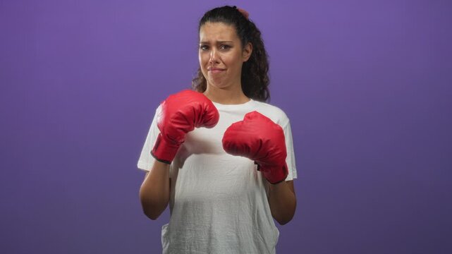 Woman wearing red boxing gloves holds guard pose in studio with purple backdrop and worried expression; nervous resilience.