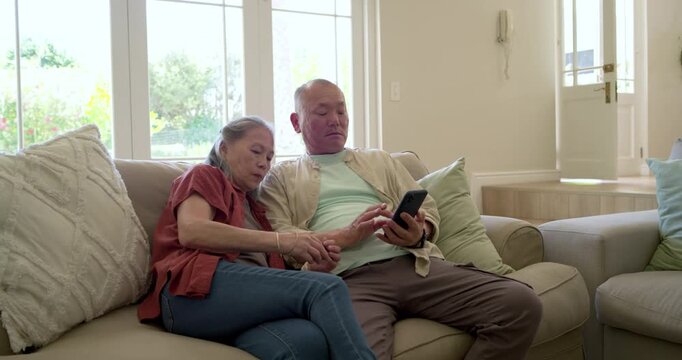 Senior Asian couple sitting on beige sofa man tapping phone woman showing credit card for payment