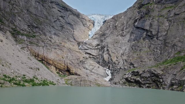 Drone view toward Briksdalsbreen glacier and lake in Norway. Meltwater flows into the basin between steep cliffs, creating a serene summer mountain scene.