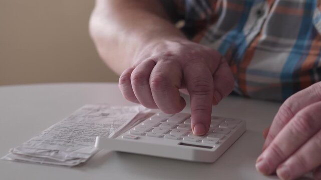 White Man Calculating Bills At Table With Calculator And Receipts, Tense Focus On Wrinkled Hands Pressing Keys, Stacks Of Receipts And Pen Nearby, Domestic Living Room Light, Slow Methodical
