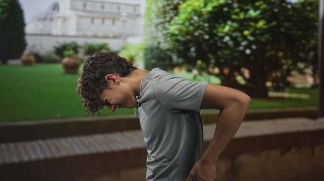 Teenage boy with hand on lower back leaning forward beside a stone building wall on a manicured garden path; discomfort.