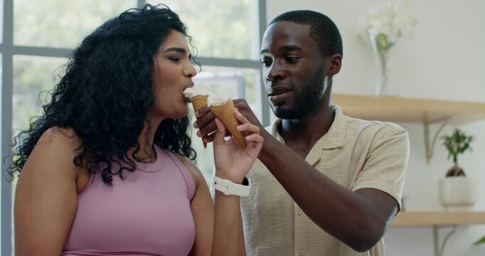 Diverse couple sharing soft-serve cones at bright window, man holding cone near woman teasing bite