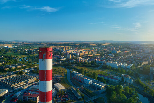 Industrial, red and white chimney rising above urban energy district with city skyline in background. Ideal for: district heating, smart city energy, urban infrastructure, decarbonization strategy.