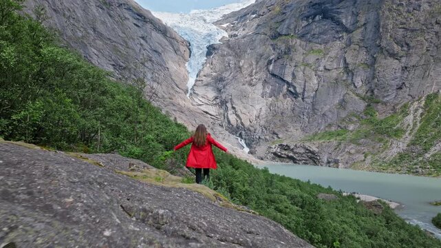 Young woman in red jacket standing near Briksdalsbreen glacier in Norway. Tourist looks toward the glacier, cliffs and milky lake on a summer day, enjoying nature, adventure travel and dramatic alpine