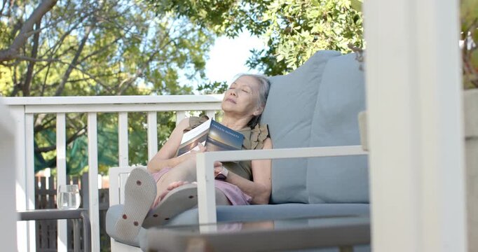 Sunlight filtering trees is lulling senior woman on porch sofa holding paperback on chest into nap