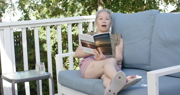 Senior Asian woman is reading open book on deck sofa, adjusting, speaking softly to relax