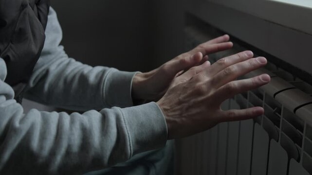 No heating at home, man warming hands near radiator in cold apartment, energy crisis lifestyle.