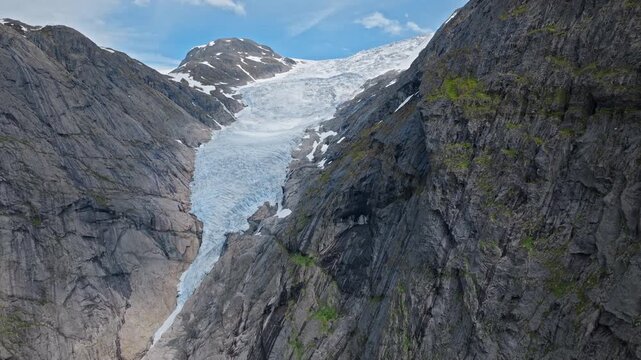 Aerial view of Briksdalsbreen glacier flowing between steep mountain cliffs in Norway. Blue ice contrasts with dark rock walls under bright summer skies, showing dramatic alpine nature.