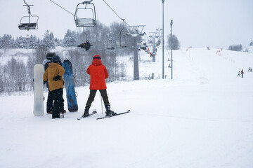 A group of two snowboarders and a skier against the backdrop of a panoramic view of a ski slope and a chairlift with passengers.