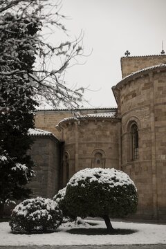 Heavy snow blankets the garden bushes outside the rear Romanesque exterior of San Pedro Church in Avila, Spain, highlighting the ancient stone architecture on a freezing winter day.