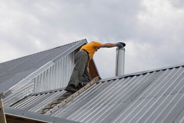A man in an orange T-shirt is climbing onto an iron roof to fix a chimney. The man on the roof is...