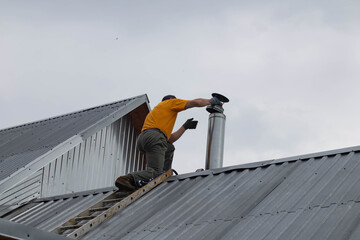 A man in an orange T-shirt is climbing onto an iron roof to fix a chimney. The man on the roof is putting a cap on the chimney.