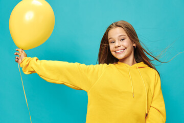 Obraz premium Smiling girl wearing yellow sweater holding yellow balloon on blue background with hair blowing and cheerful expression for happiness and celebration concept.