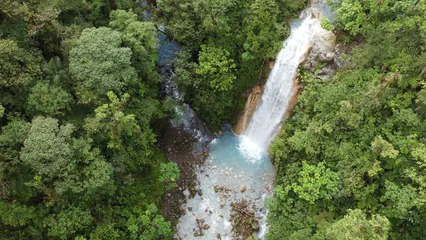 Fototapete Wald Fluss High-angle drone shot of a tropical waterfall flowing into a bright blue river pool  © Jose